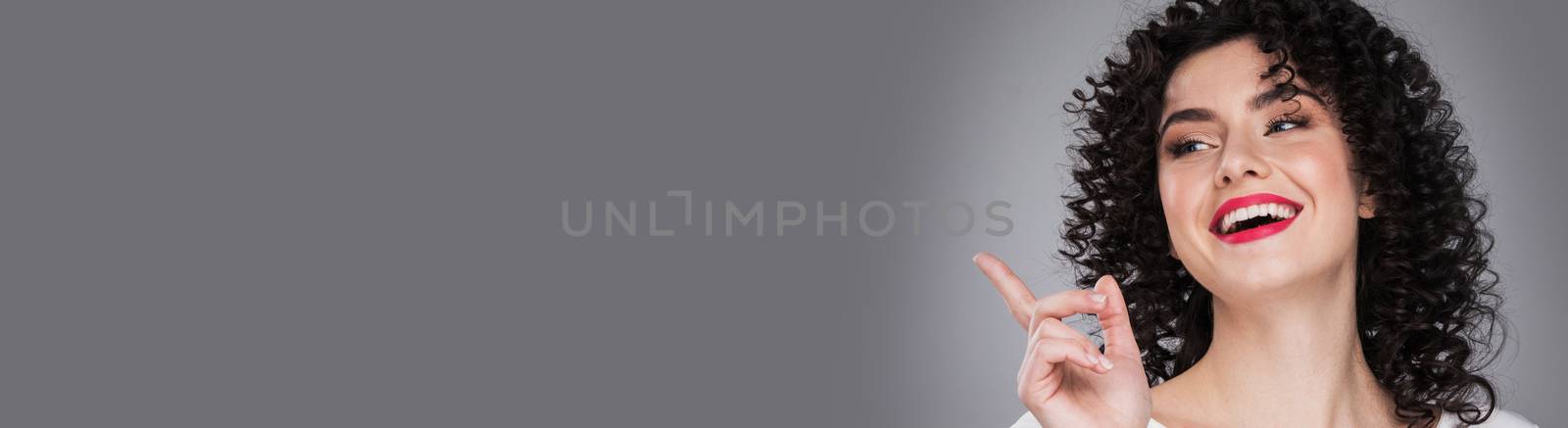 Woman showing product .Beautiful girl with curly hair pointing to the side . Presenting product. Gray background with copy space