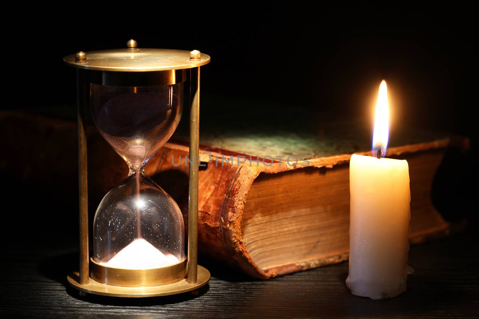 Vintage hourglass near lighting candle on dark background