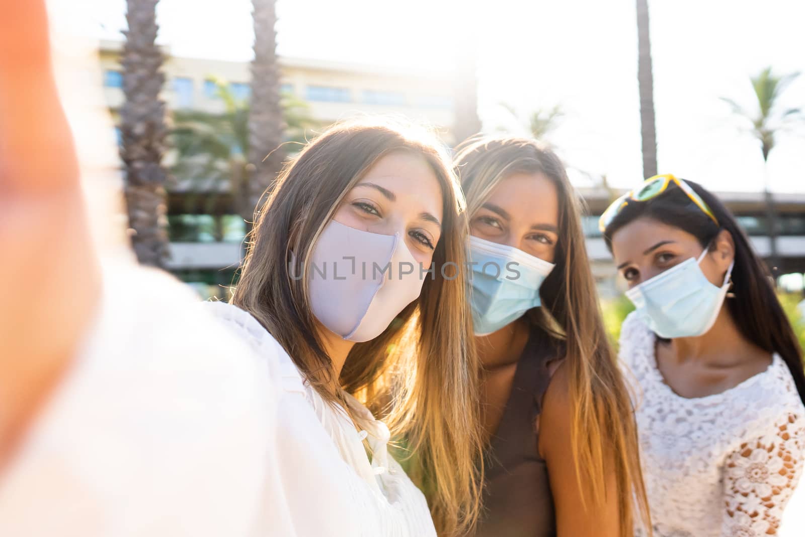 Cute blonde young woman take a selfie with her two female friends wearing the Coronavirus protection mask - New normal life with Covid-19 pandemic using social network connections by robbyfontanesi