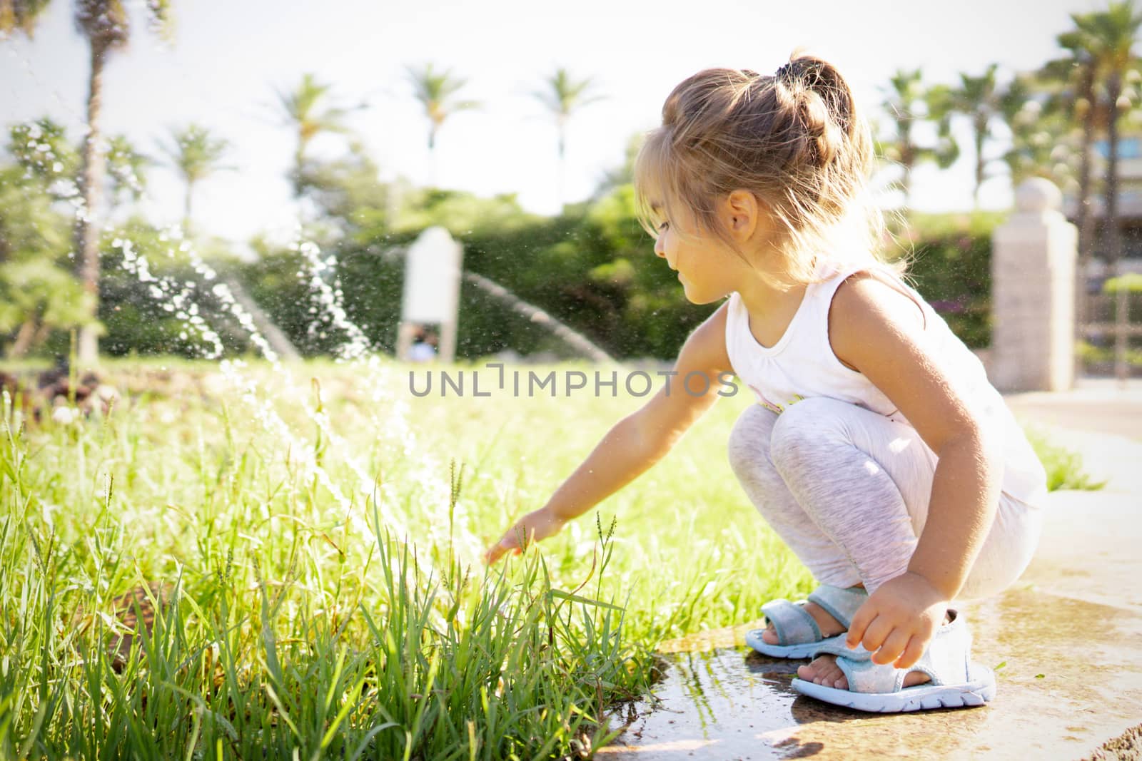 Female blonde cute child bent over the edge of a fountain in the park, touching the water jets with her hand - Concept: the importance and value of water for the future of the planet and our children