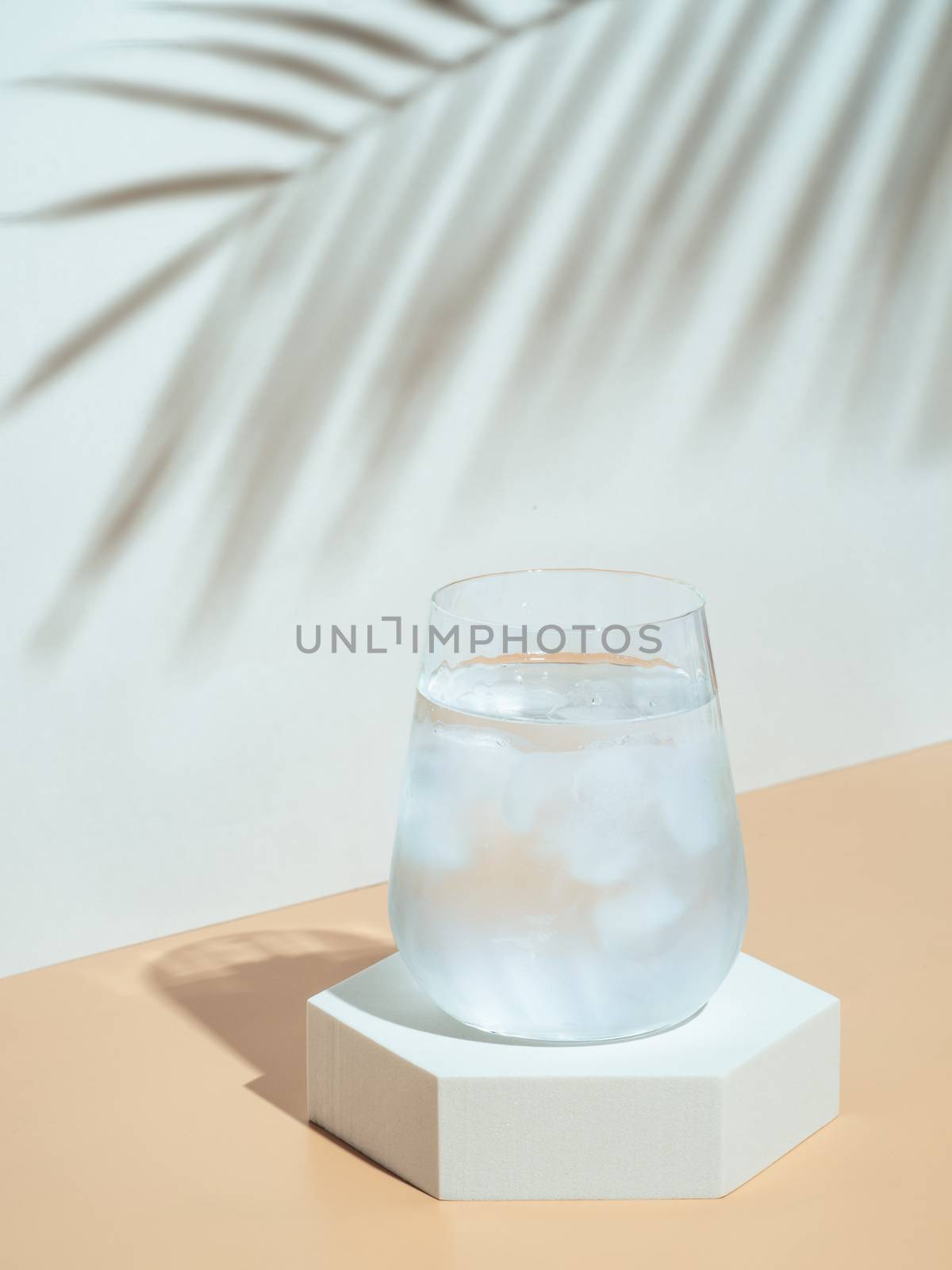 Cold water with ice in tumbler glass on hexagon pedestal. Mockup for drink in fashion trendy style with tropical leaf shadow. Modern still life with glass. Copy space. Health, beverage, summer concept