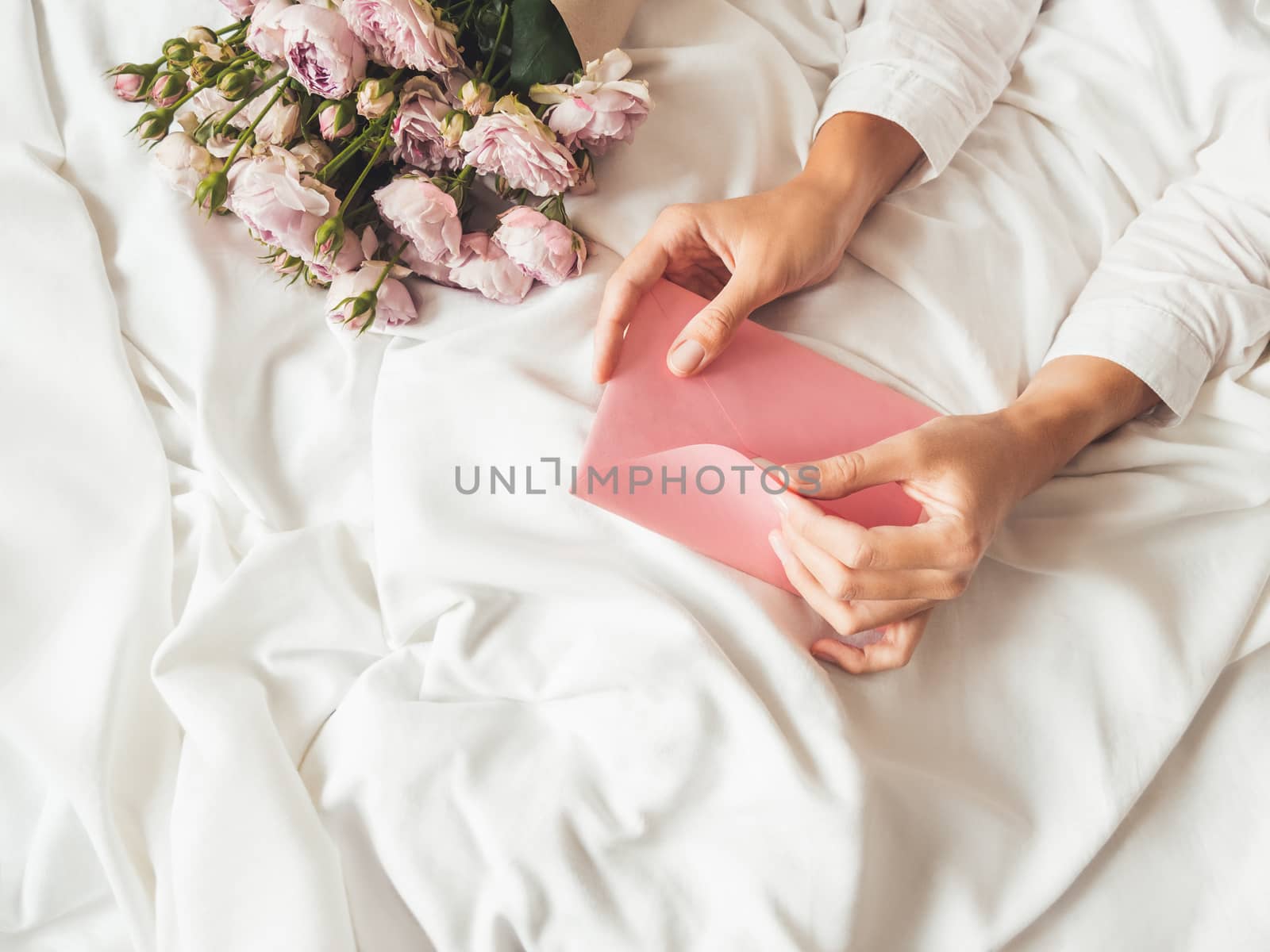 Woman is lying on crumpled bed sheet. She received bouquet of lilac colored roses and letter in pink envelope. Cute present for Valentine Day or Happy Birthday surprise.