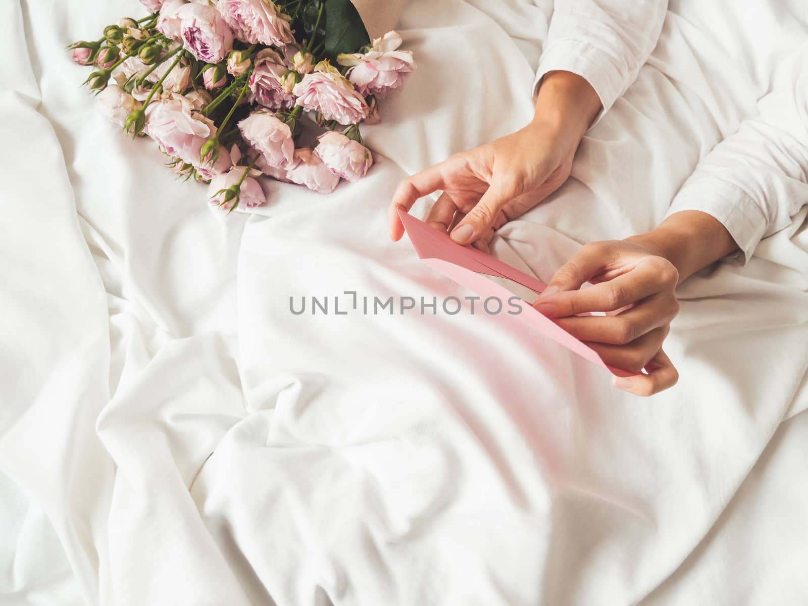 Woman is lying on crumpled bed sheet. She received bouquet of lilac colored roses and letter in pink envelope. Cute present for Valentine Day or Happy Birthday surprise.