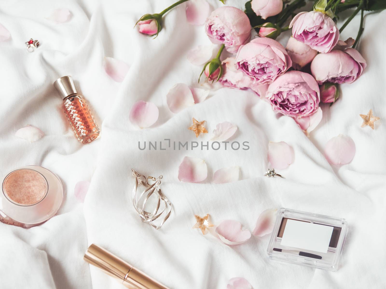 Top view on pink roses and petals on crumpled white fabric background. Decorative cosmetic - perfume, mascara, eye and brow shadows. Cute and fashionable jewelry accessories.