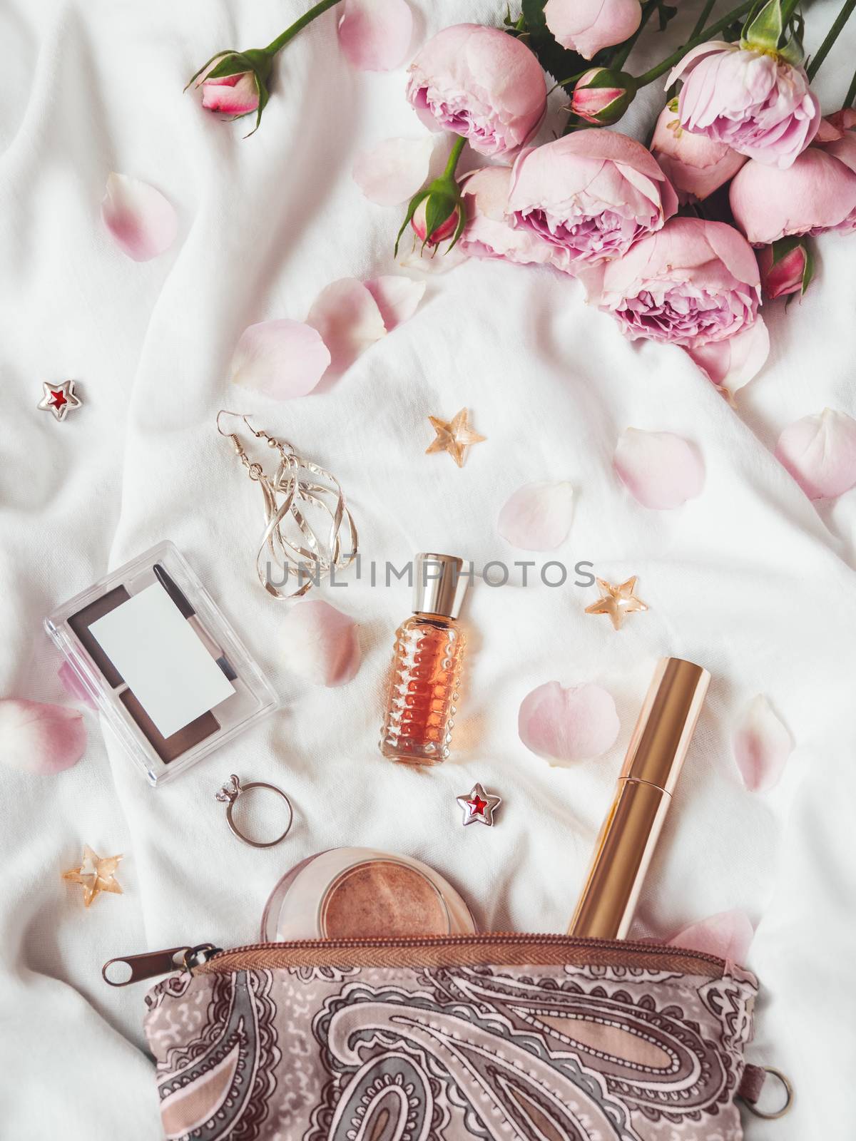 Top view on cosmetic bag on crumpled white fabric background. Decorative cosmetic - perfume, mascara, eye and brow shadows. Cute and fashionable jewelry accessories. Pink roses and petals.