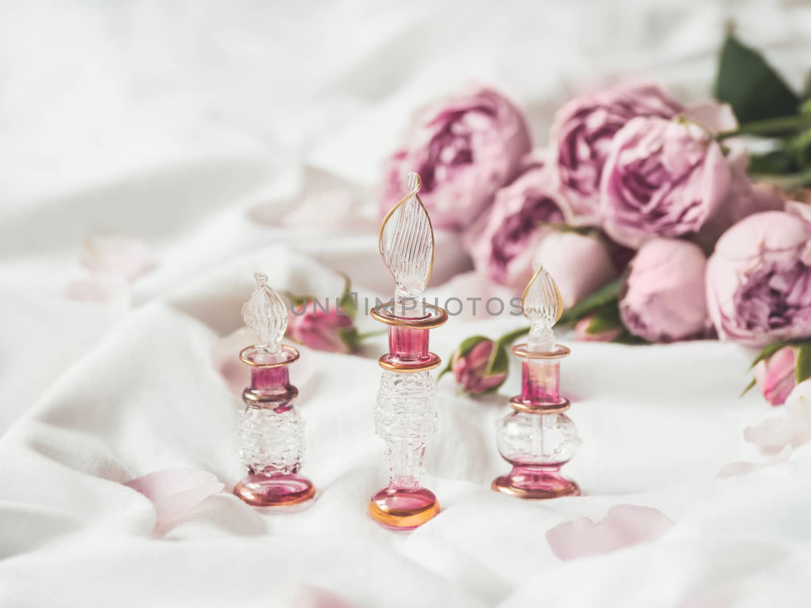 Three graceful bottles for perfume or essential oil on white crumpled fabric. Pink glass bottles with eastern ornament. Pink rose bouquet and petals as decoration.