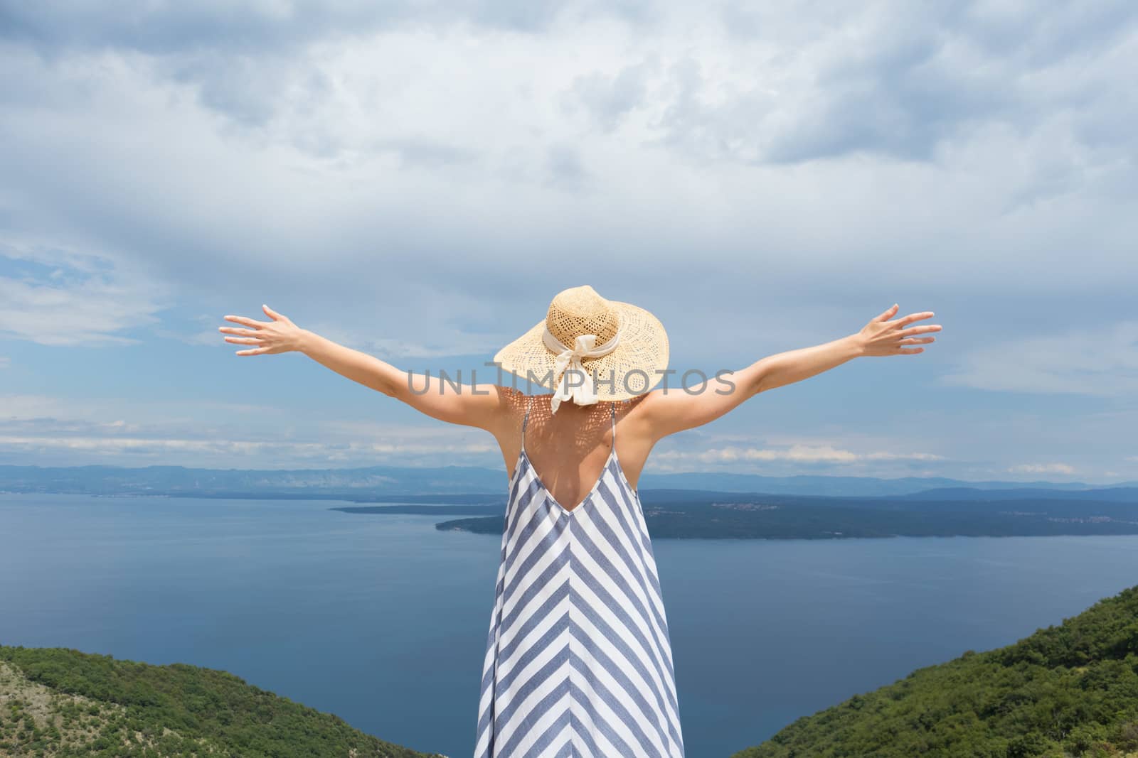 Rear view of young woman wearing striped summer dress and straw hat standing in super bloom of wildflowers, relaxing with hands up to the sky, enjoing beautiful view of Adriatic sea nature, Croatia by kasto