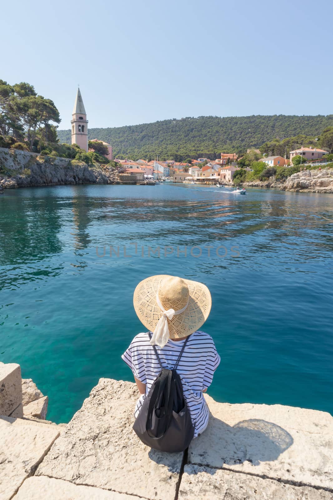 Woman traveler wearing straw summer hat and backpack, sittingat edge of stone pier, enjoying beautiful panoramic view of Veli Losinj, Losinj island, Croatia.