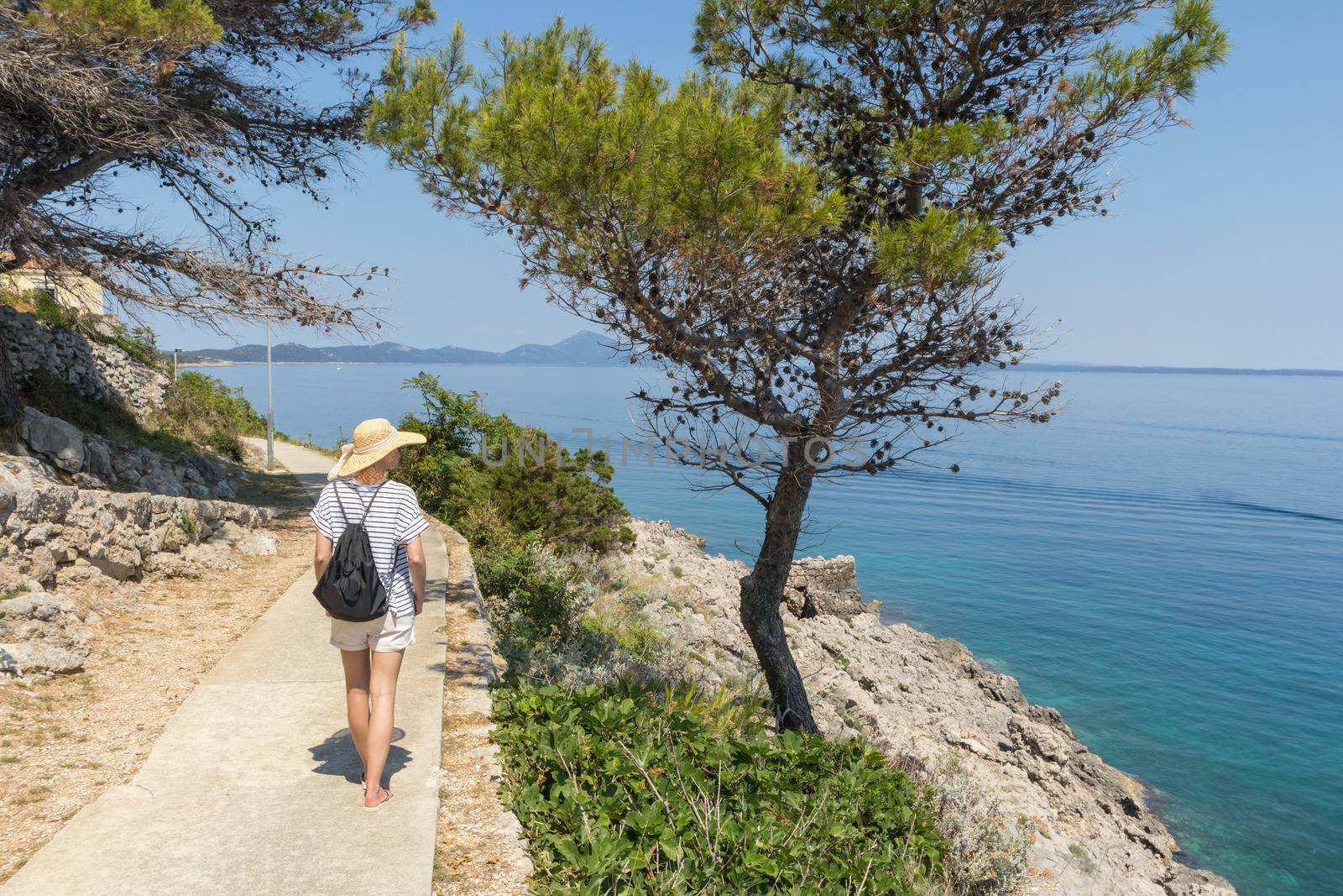 Young active feamle tourist wearing small backpack walking on coastal path among pine trees looking for remote cove to swim alone in peace on seaside in Croatia. Travel and adventure concept by kasto