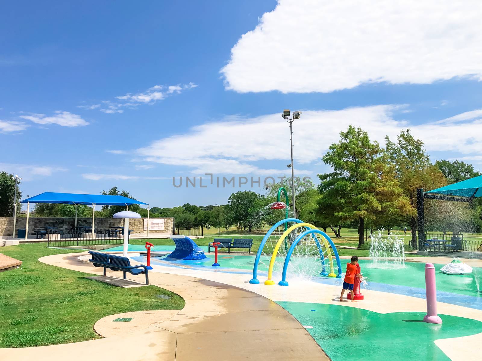 Rear view Asian toddler boy playing at splash park near Dallas, Texas, America. Colorful recreation site with splashing water fountains for kids summertime activities