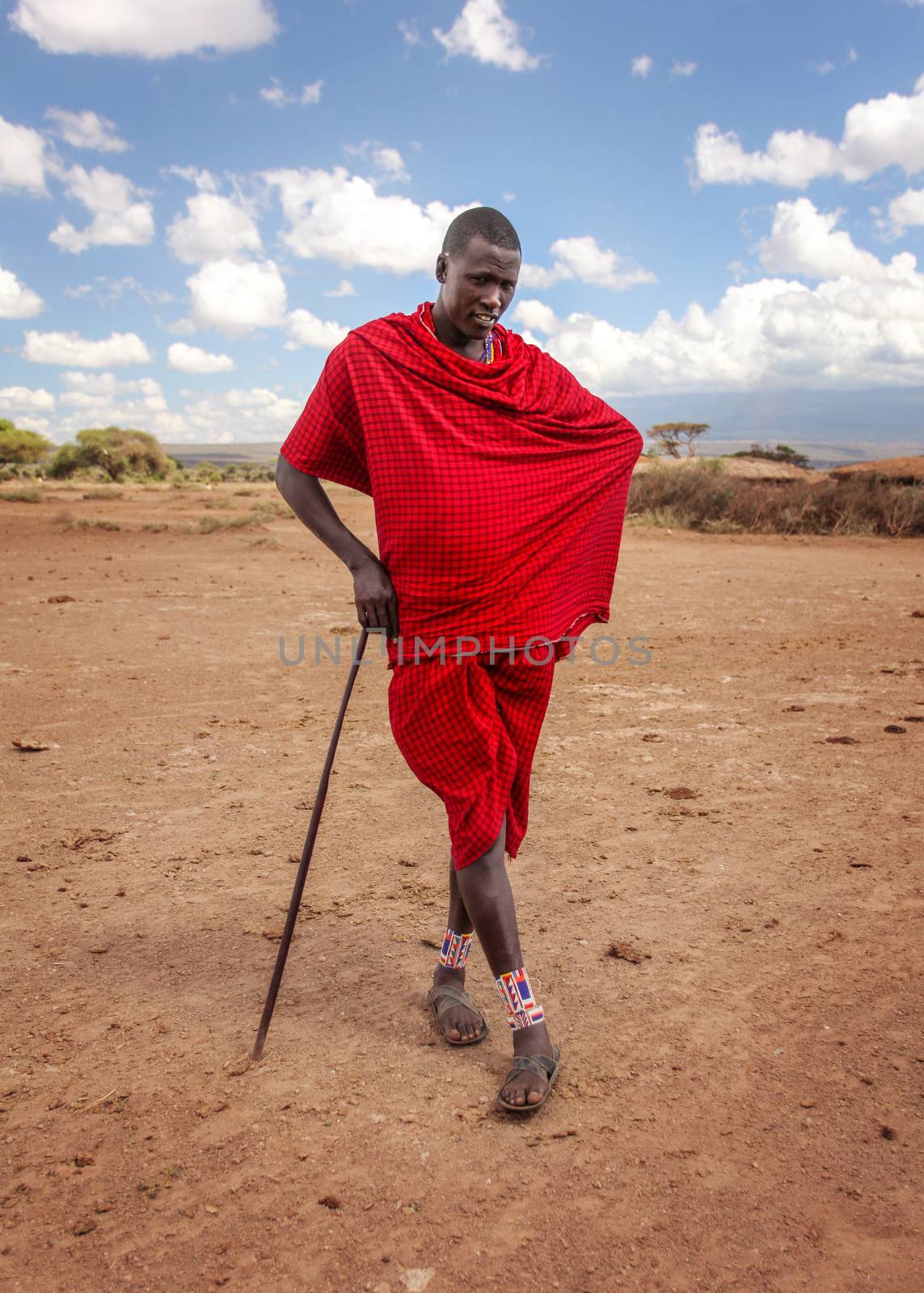 Unknown village near Amboselli park, Kenya - April 02, 2015: Unknown Masai warrior posing for tourists in traditional bright red robe, leaning on his stick.