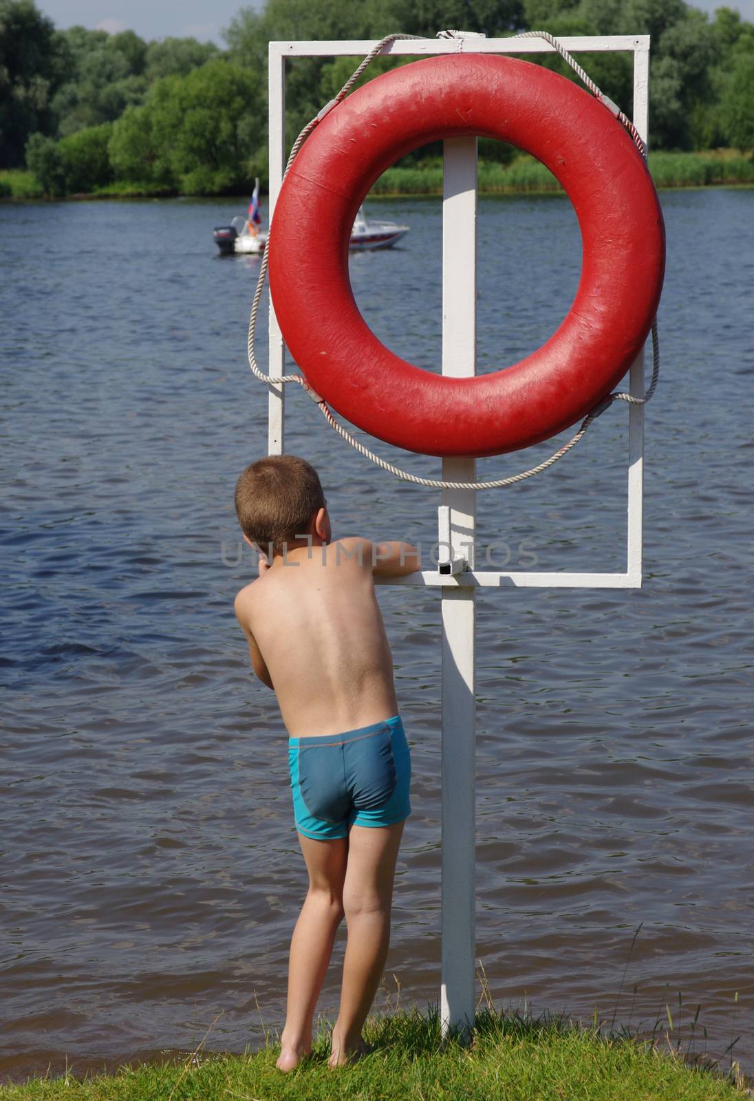 The boy is standing on the shore of the pond next to the big red life ring
