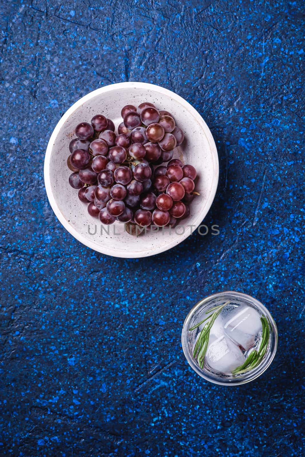 Fresh ice cold carbonated water in glass with rosemary leaf near to wooden bowl with grape berries, blue textured background, top view