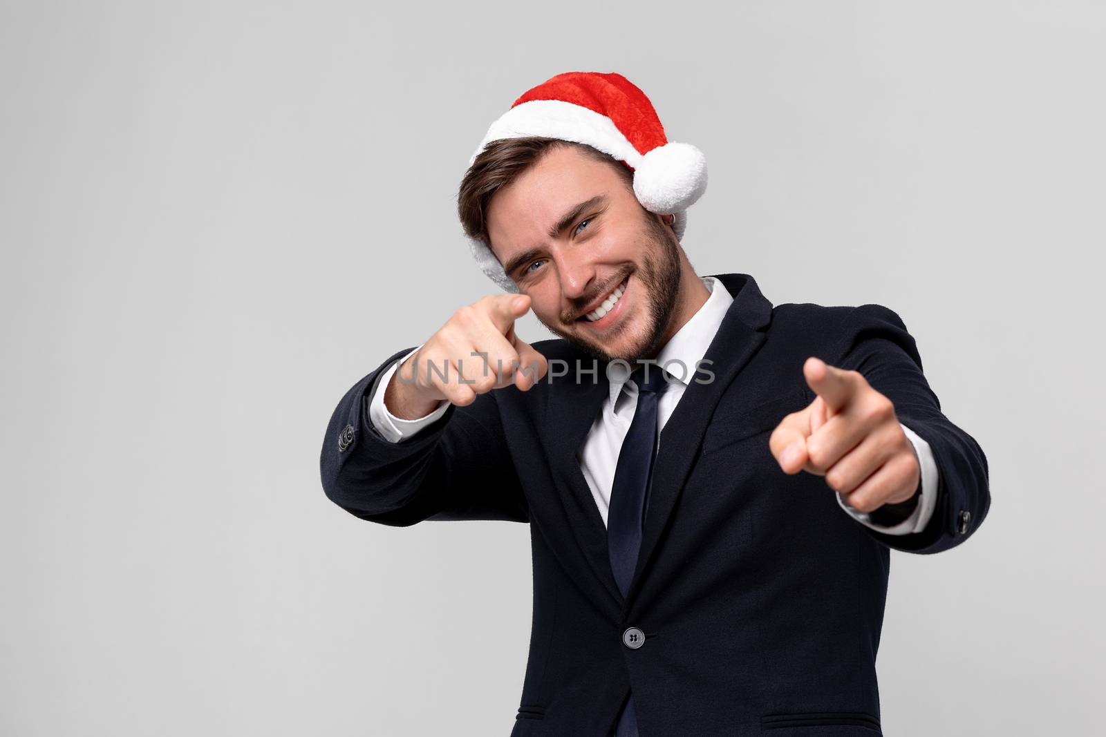 Young handsome caucasian guy in business suit and Santa hats on white background in studio smilie and showing Finger to the camera. Close up portrait business person with Christmas mood Holiday banner