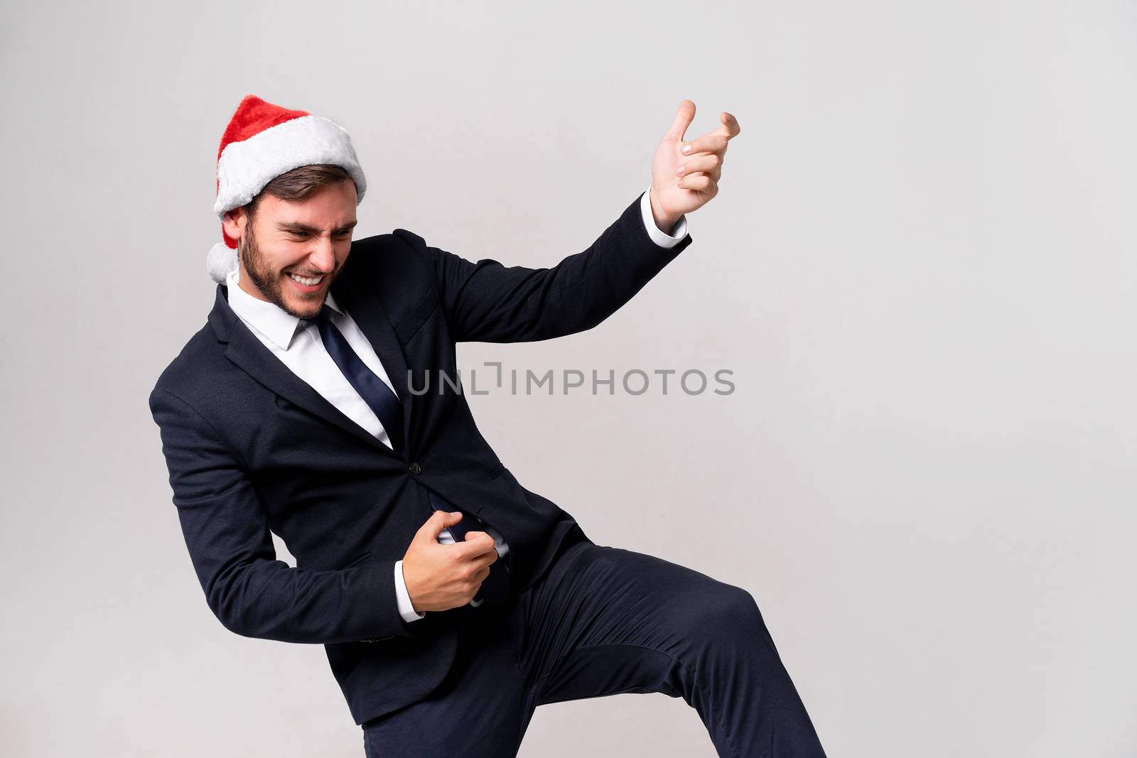 Young handsome caucasian guy in business suit and Santa hats stands on white background in studio Plays on an imaginary guitar. Close up portrait business person with Christmas mood Holiday banner