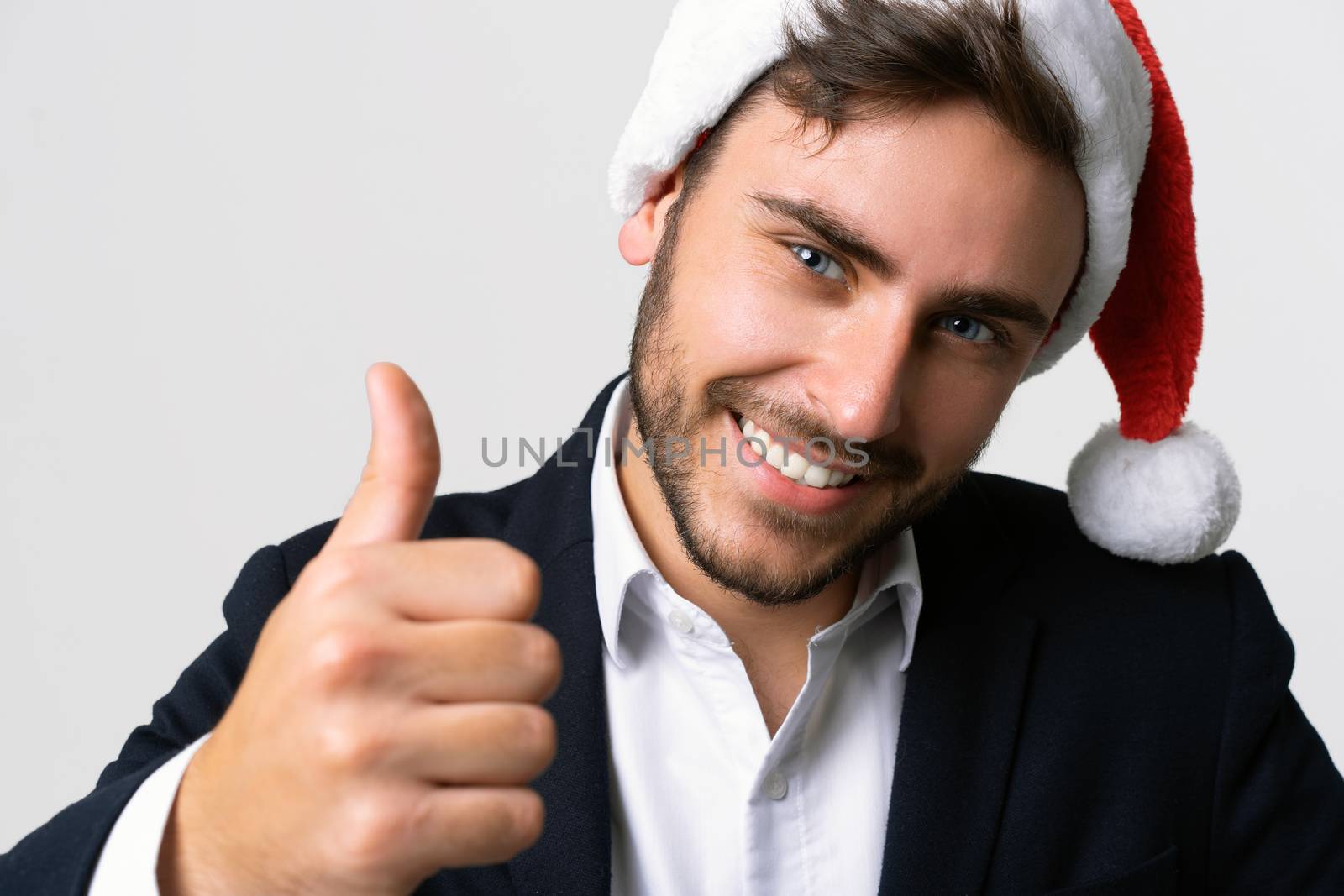 Young handsome caucasian guy in business suit and Santa hats stands on white background in studio smilie and showing thumbs up. Close up portrait business person with Christmas mood Holiday banner