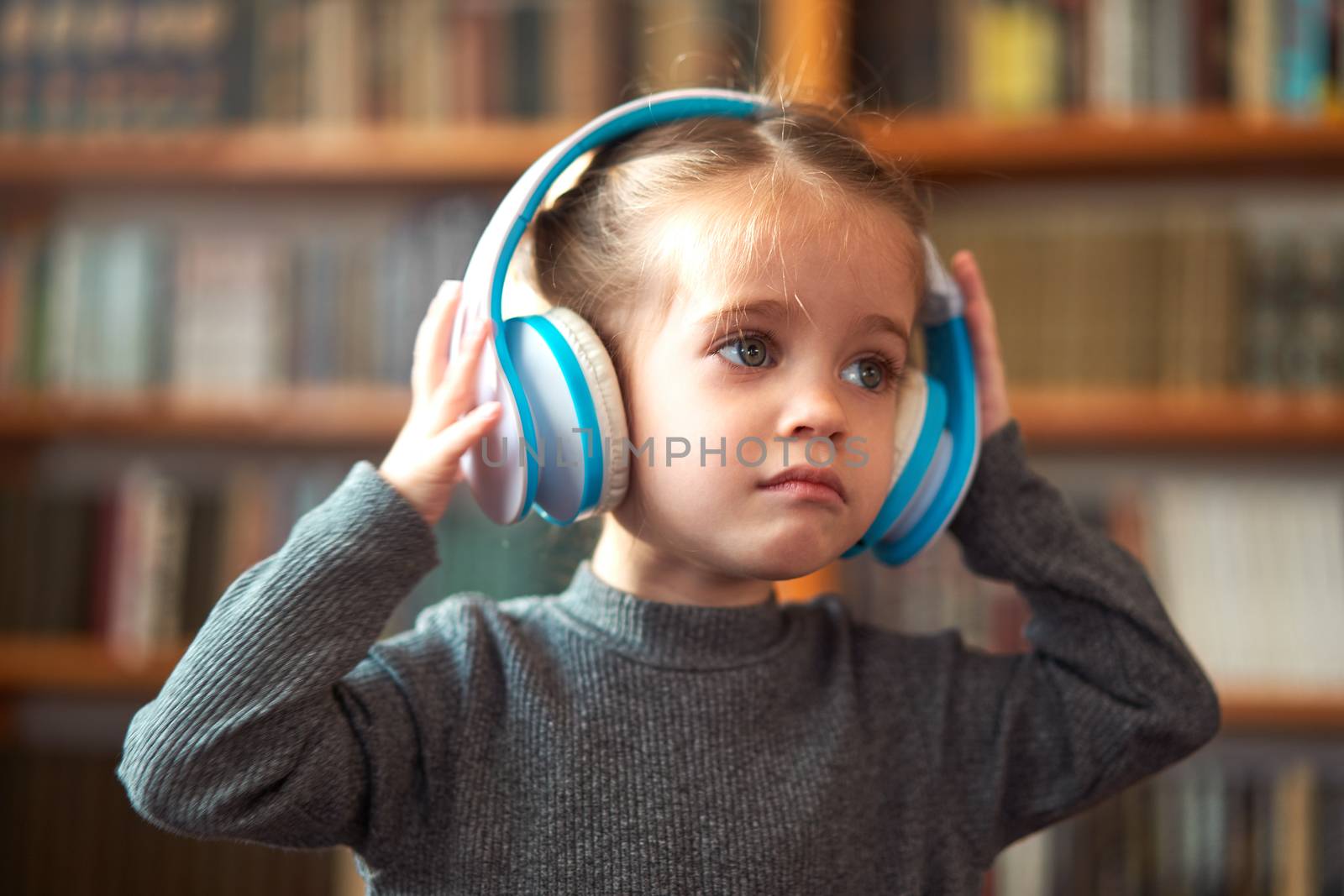 Cute Caucasian little girl in big white headphones listens music in the interior against the background of bookshelves Modern children. Happy childhood with technology Child spends free time at home.
