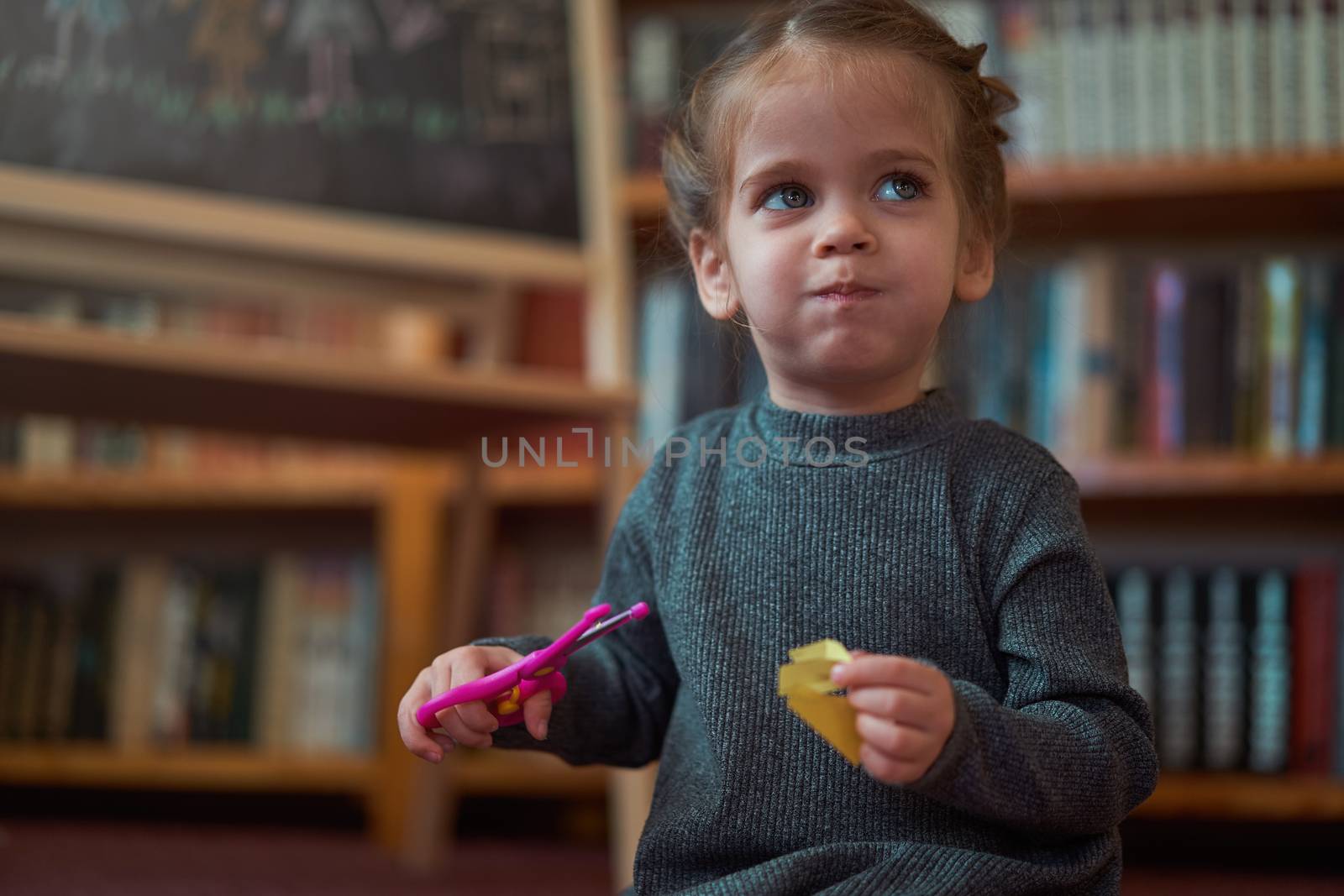 Cute little caucasian girl is playing with scissors and colored paper while sitting on the floor at home. Handcraft for little children. Home leisure