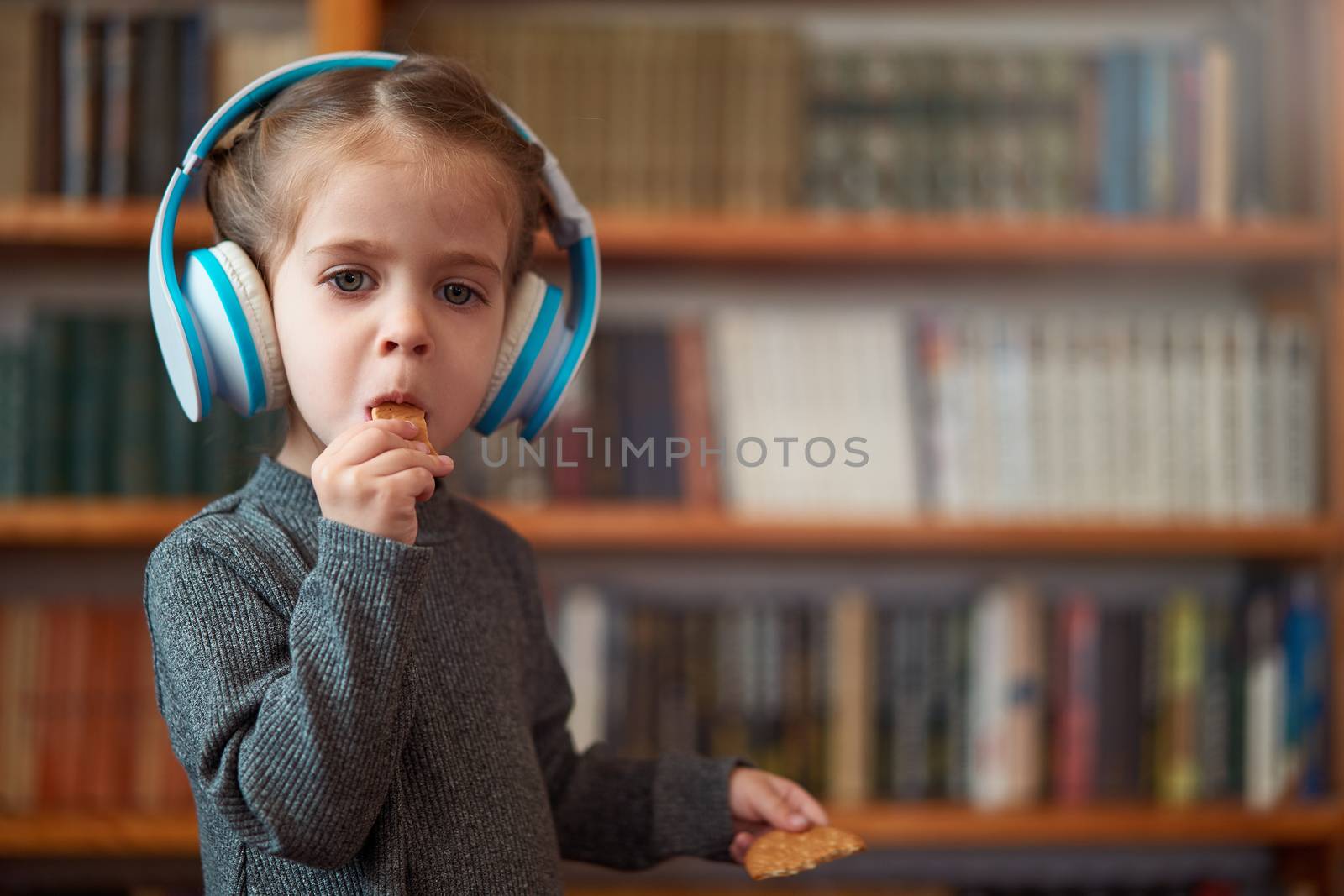 Cute Caucasian little girl in big white headphones listens music in the interior against the background of bookshelves Modern children. Happy childhood with technology Child spends free time at home.