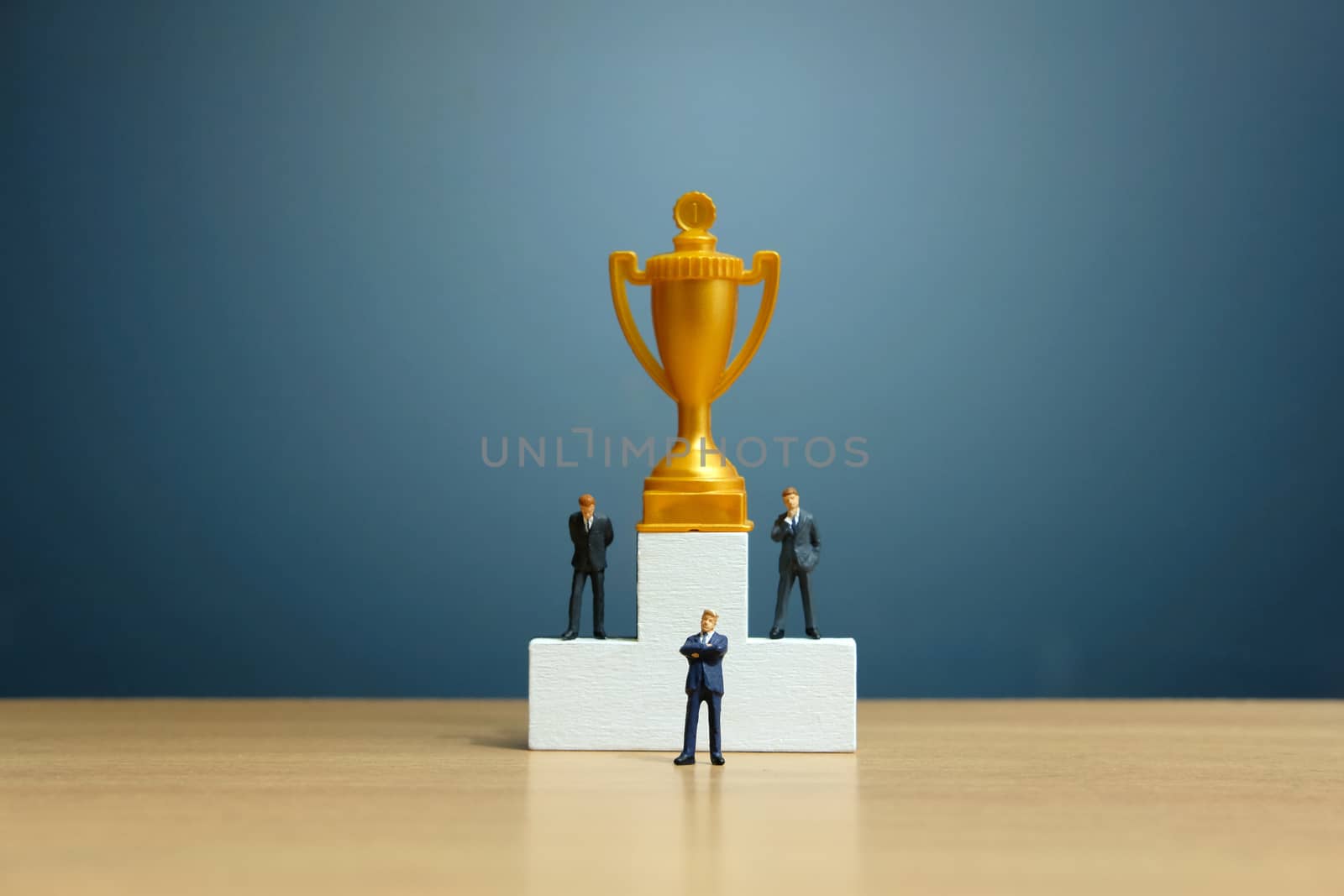 Miniature business concept - businessman standing in front of white winner podium with golden trophy