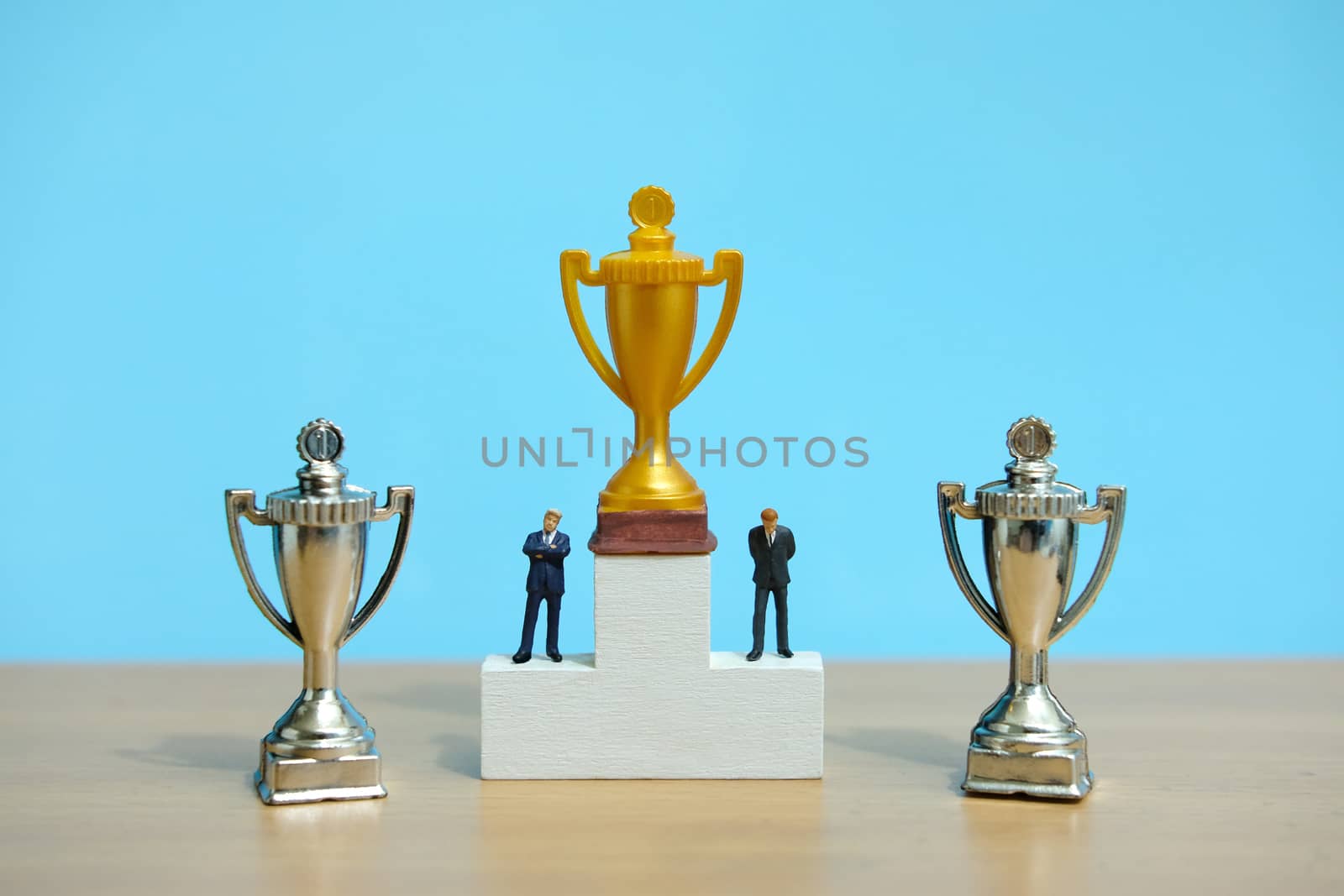 Miniature business concept - businessman standing on white winner podium with golden and silver trophy