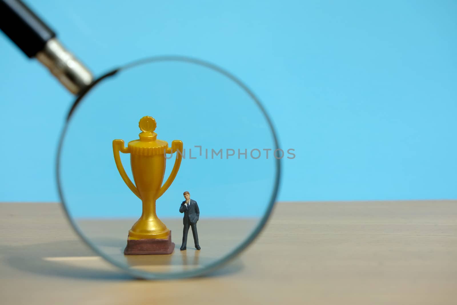 Miniature business concept - businessman standing in front of golden trophy and magnifier glass