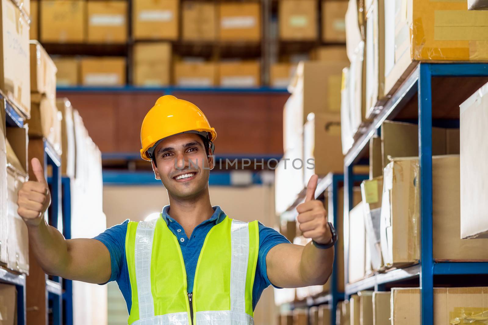Portrait of Indian warehouse worker man with safety clothes standing with confident and showing thumps up in local workplace with warehouse or factory, success and achievement concept.