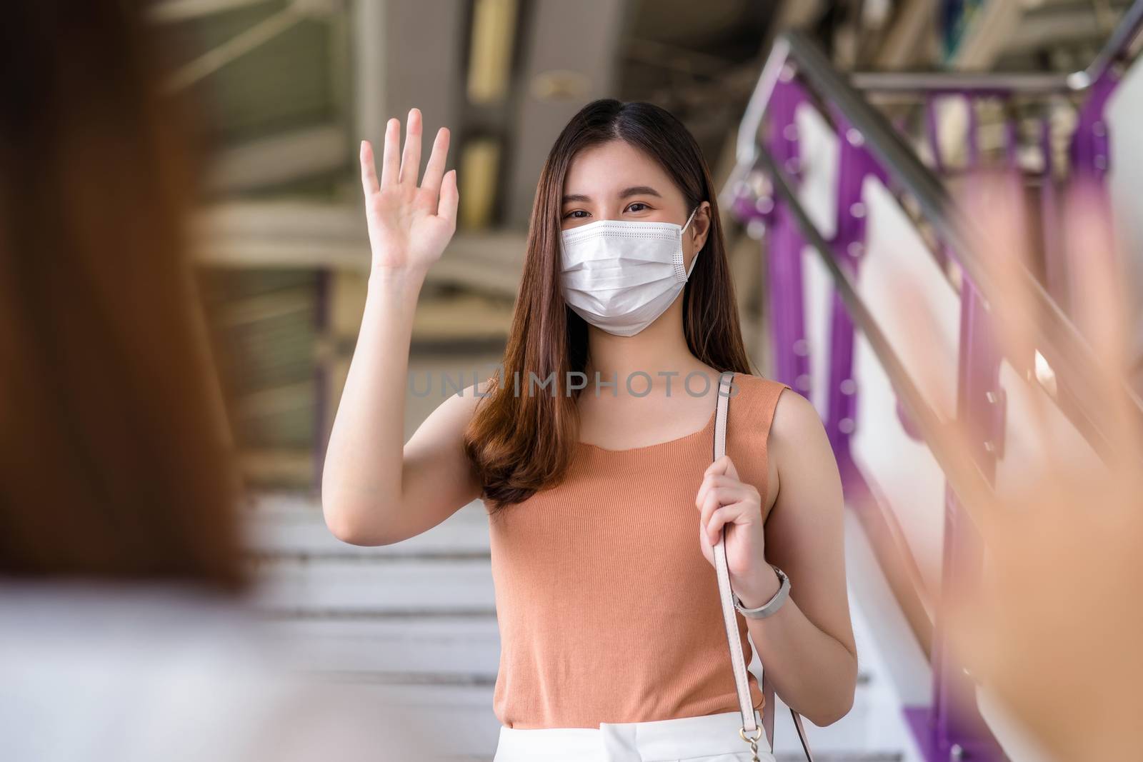 Young Asian woman passenger wearing surgical mask and Waving hand for greeting to her friend in subway train when traveling in big city at Covid19 outbreak, social distancing and new normal concept 
