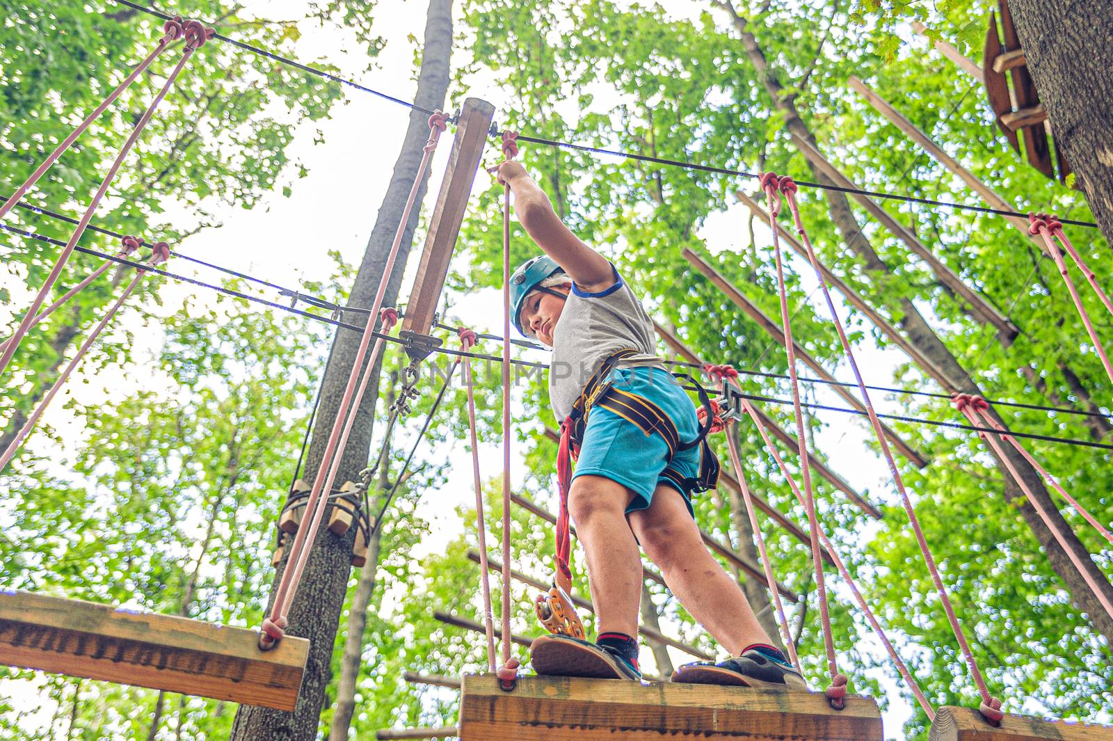 Teen boy in gear and helmet holds on to the ropes in forest adventure park by chernobrovin