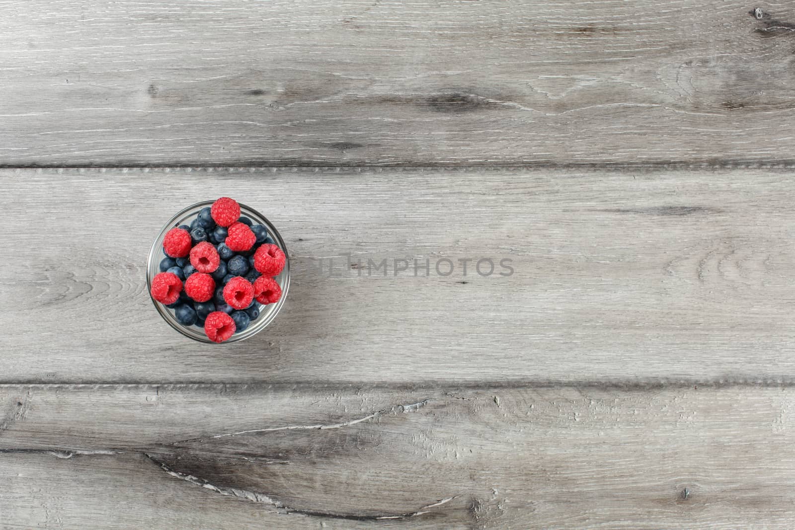 Table top view on small glass bowl full of blueberries and raspberries on gray wood table.