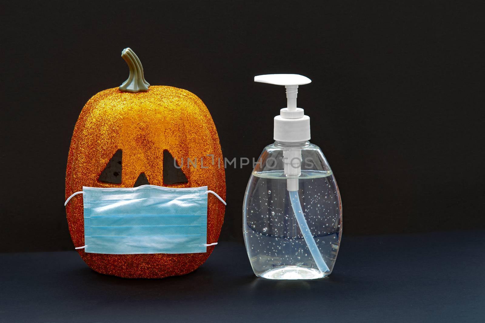 Jack-o-lantern pumpkin Halloween wearing a face mask with hand sanitizer bottle on a black background. Concept: Halloween during global pandemic. Covid-19, Coronavirus. by oasisamuel