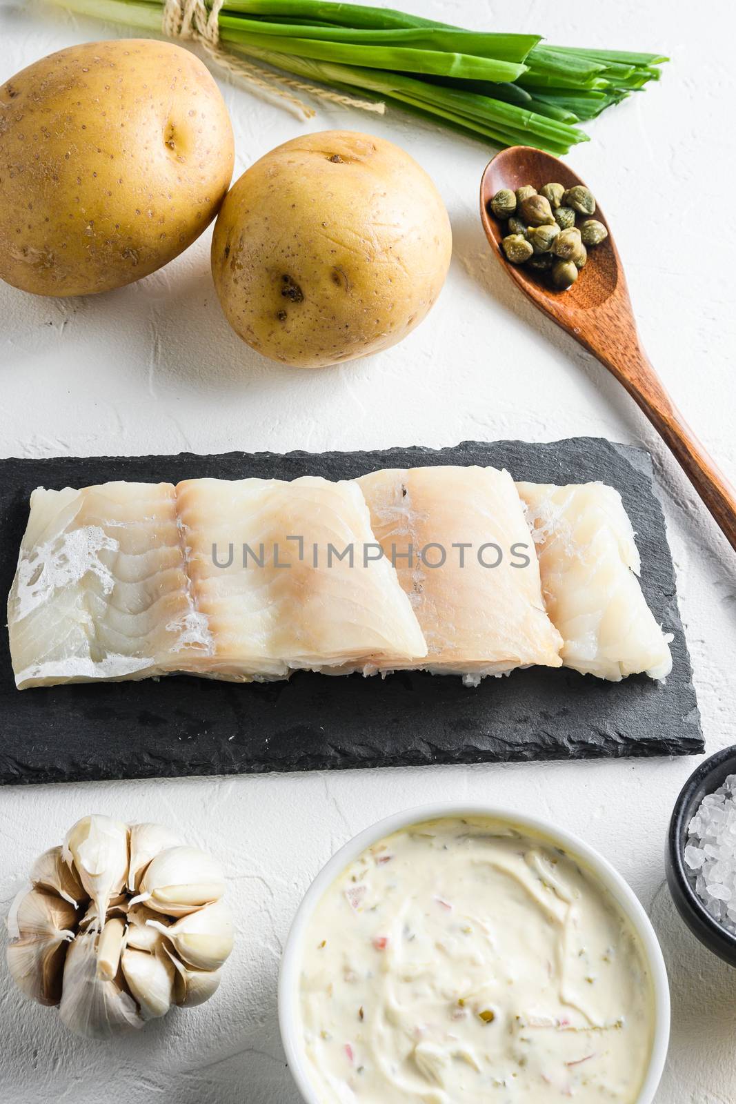 Traditional fish and chips ingredients recipe raw cod fillets on stone slate batter, potatoe, tartar sauce, lemon, capers , green herbs garlic, salt, peppercorns on white stone background side view close up vertical.