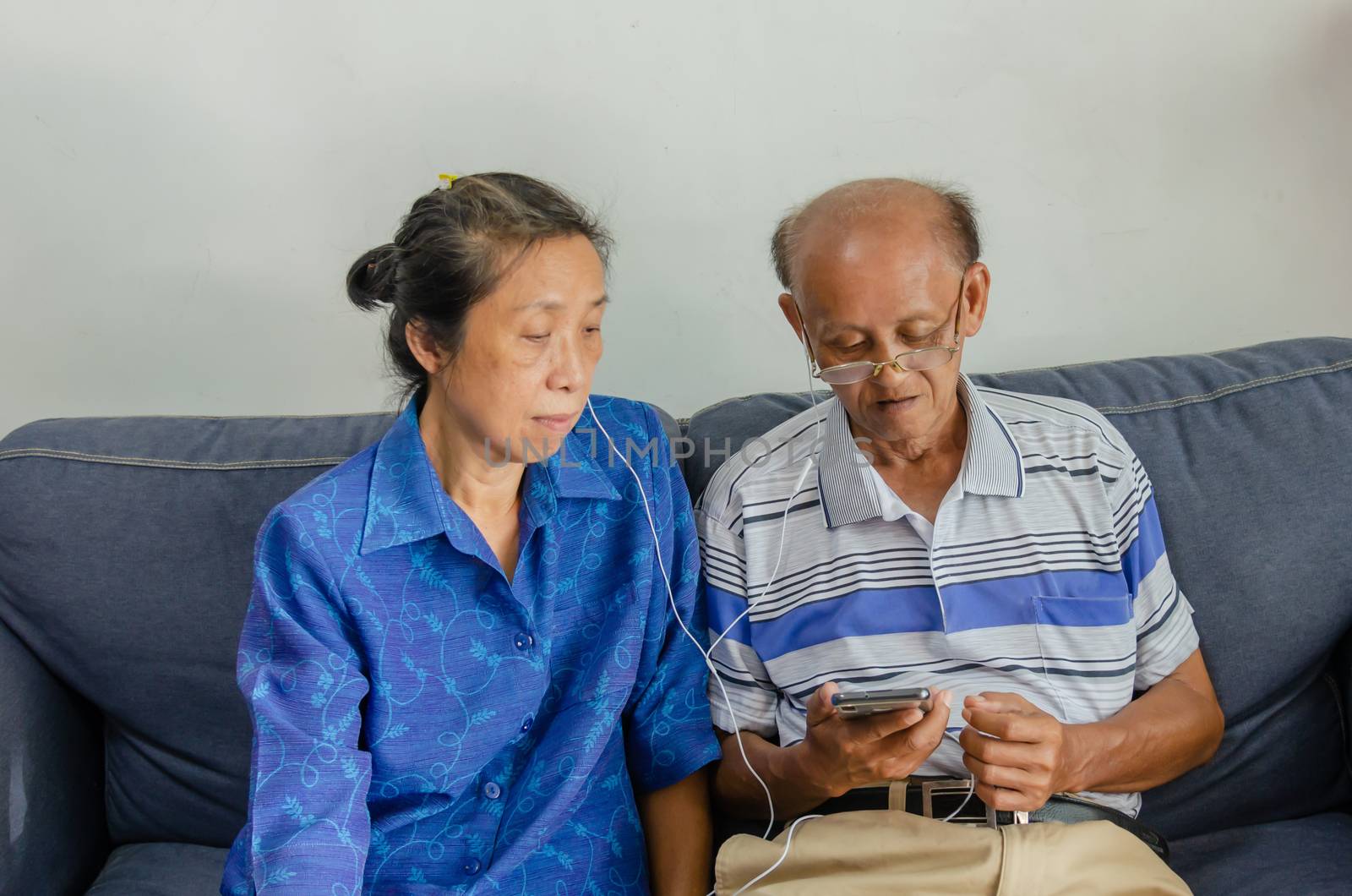 Asian elderly couples watch mobile phones and use headphones on the sofa.