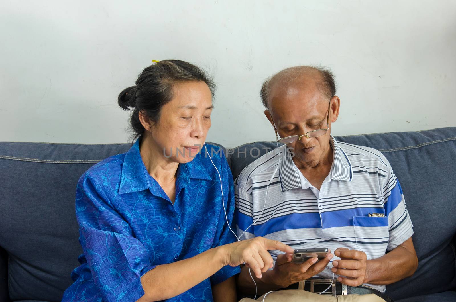 Asian elderly couples watch mobile phones and use headphones on the sofa.