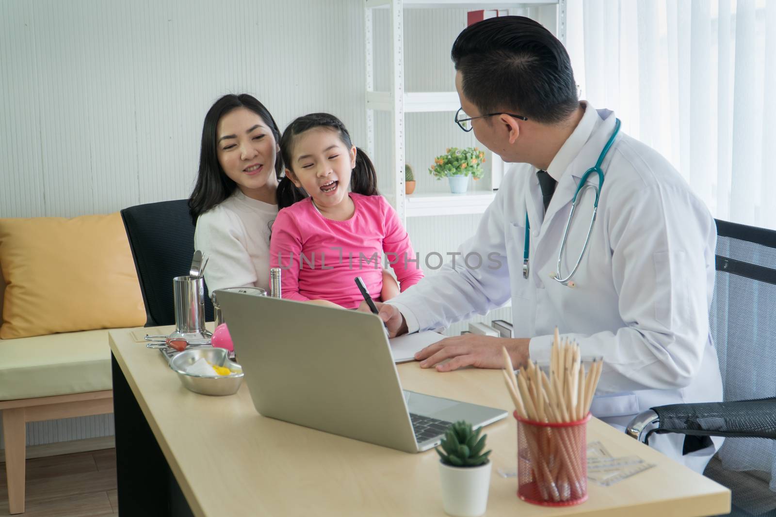 Happy Asian adult mother and preschool daughter consult pediatrician at the clinic. Young Asian male doctor examines physical health and asks for information from girls' parent