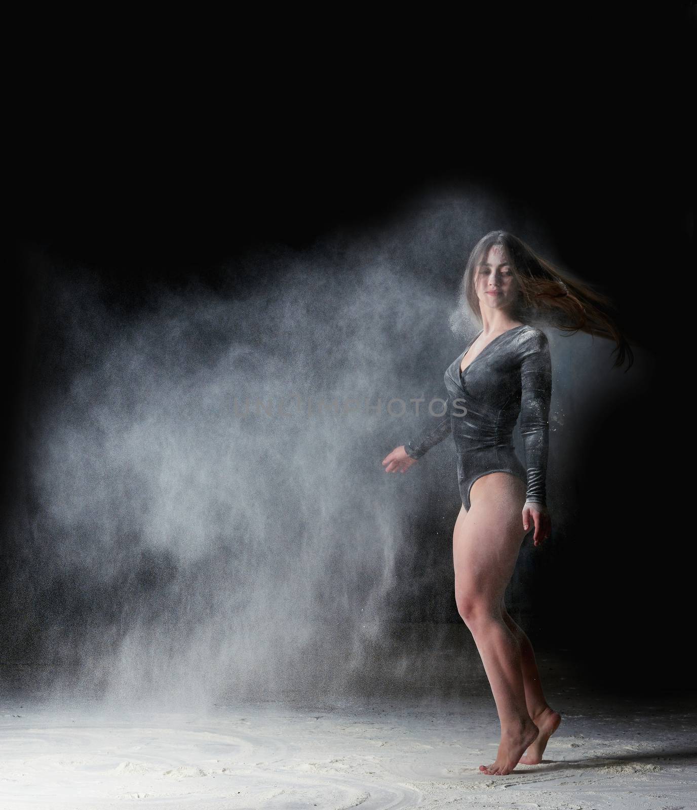 beautiful young caucasian woman in a black bodysuit with a sports figure is dancing in a white cloud of flour on a black background