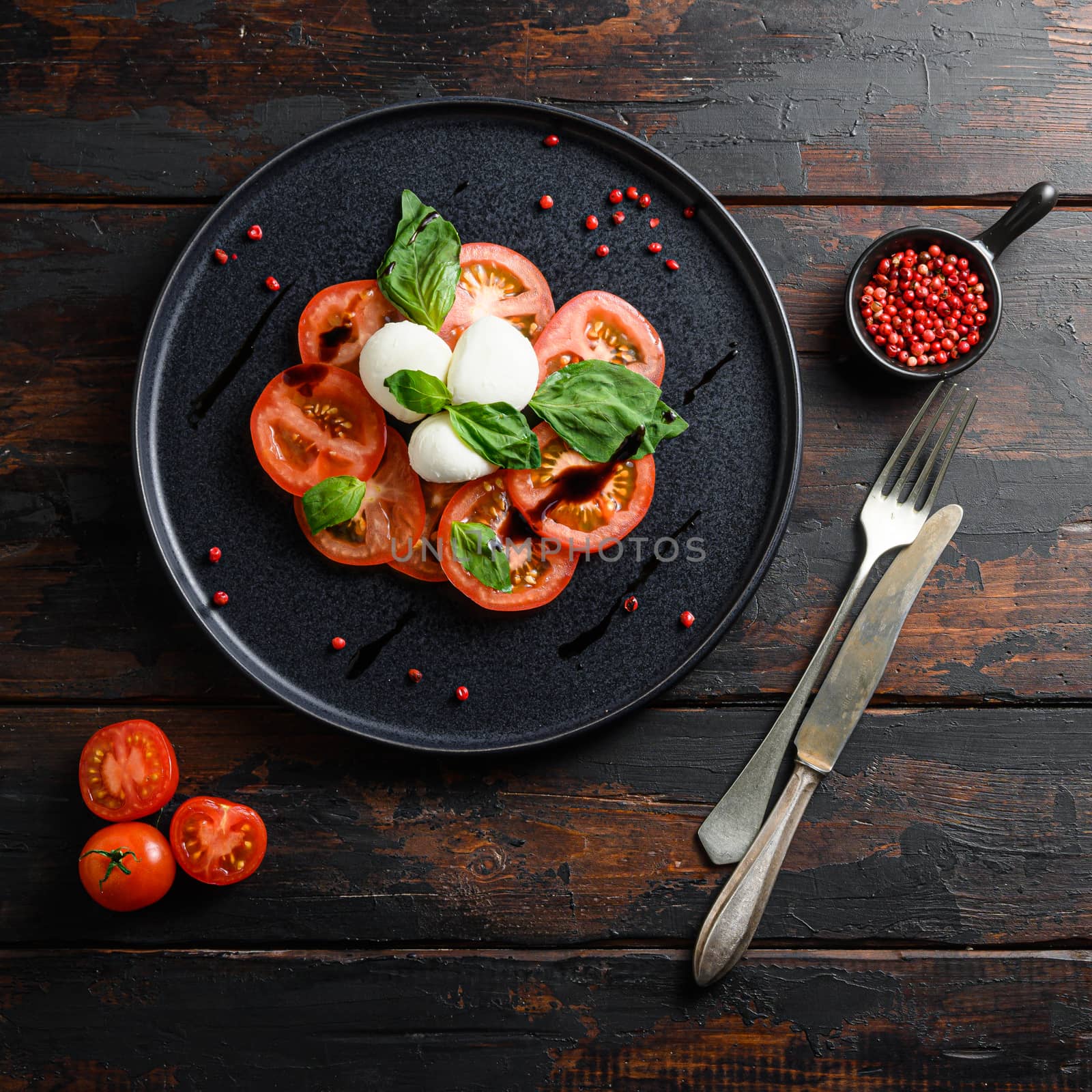 Caprese salad Tomato and mozzarella slices with basil leaves on olive wood dark table top view on black plate square crop.