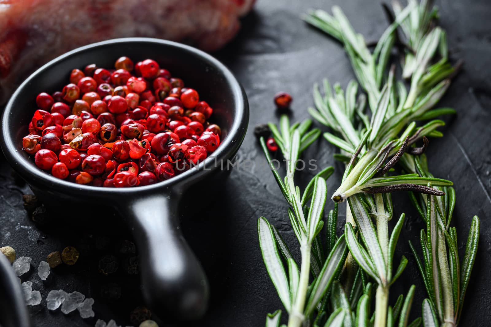 Rose Peppercorn and Rosemary herbs close up on black stone table with spices and raw meat near side view selective focus.