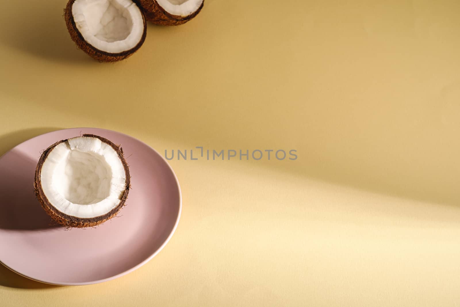 Coconut half in pink plate with nut fruits on cream yellow plain background, abstract food tropical concept, angle view copy space