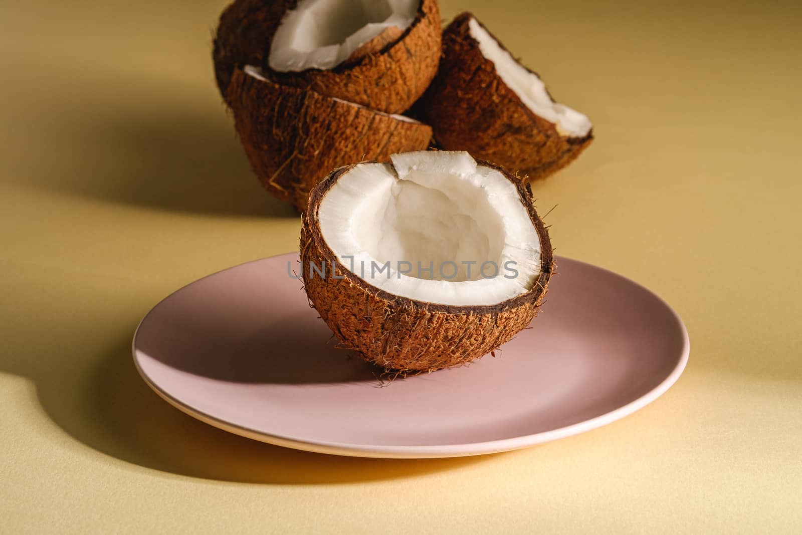 Coconut half in pink plate with nut fruits on cream yellow plain background, abstract food tropical concept, angle view