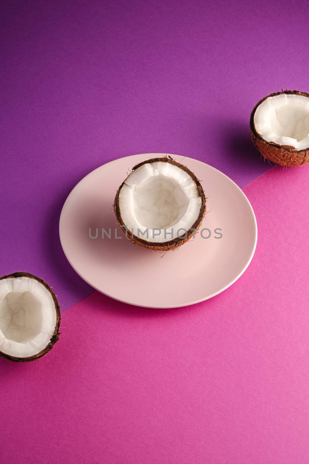 Coconut half in pink plate with nut fruits on violet and purple plain background, abstract food tropical concept, angle view