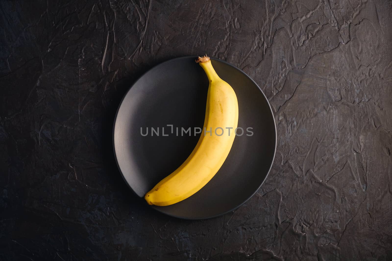 Single banana fruit in black plate on dark textured background, top view