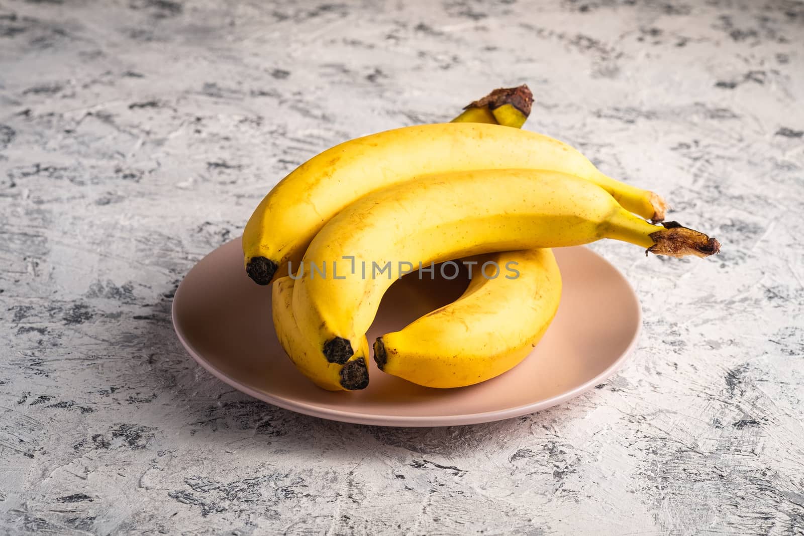Banana fruits in pink plate on white textured background, angle view copy space