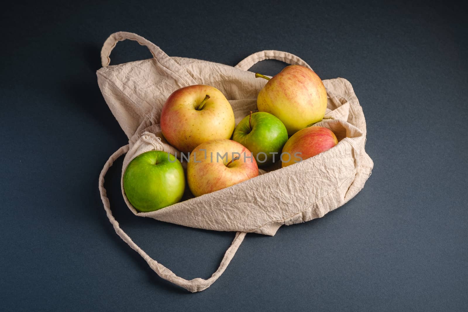 Fresh sweet apples in reusable textile grocery bag on dark grey background, angle view