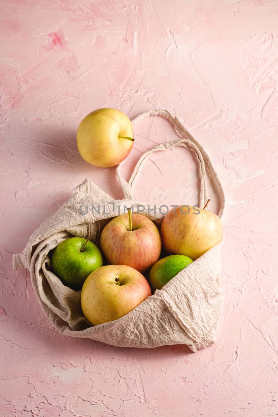 Fresh sweet apples in reusable textile grocery bag on pink textured background, angle view