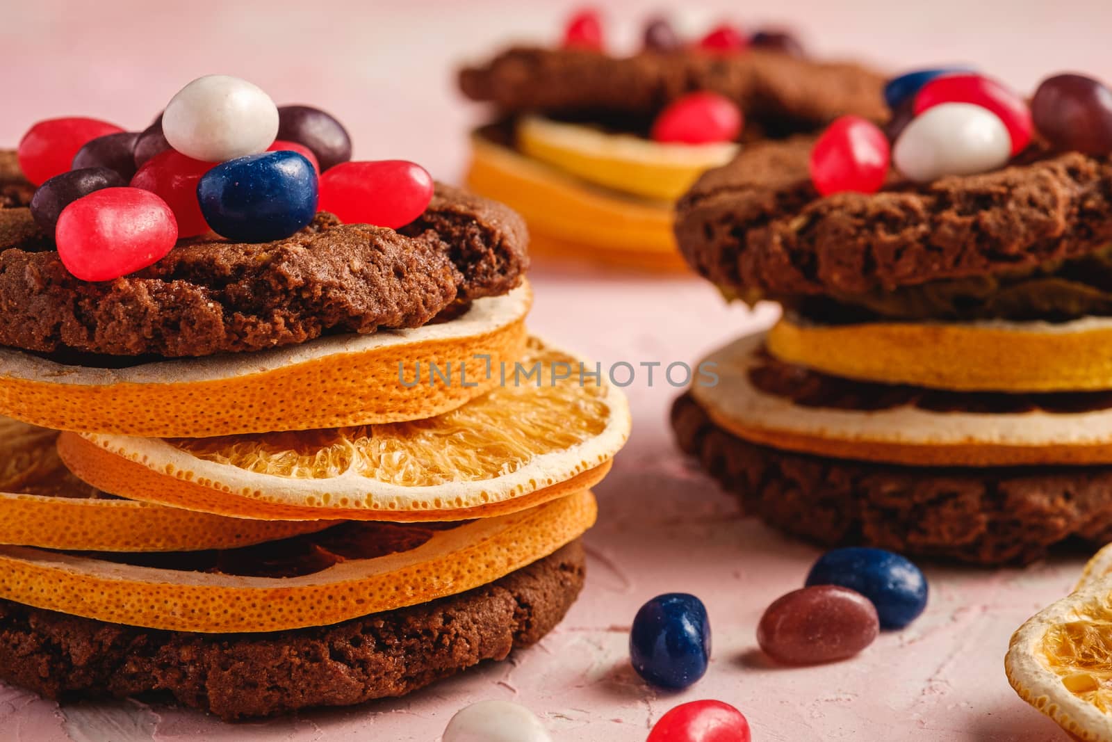 Homemade oat chocolate cookies sandwich with dried citrus fruits and juicy jelly beans on textured pink background, angle view macro