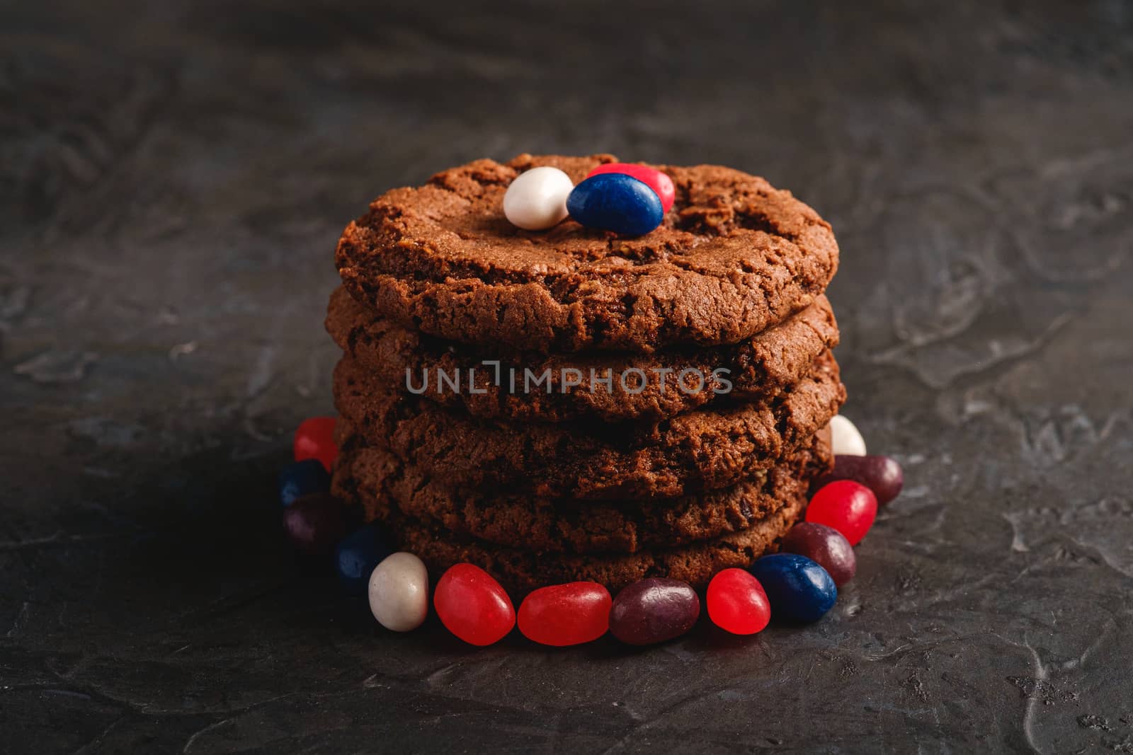 Homemade oat chocolate cookies stack with cereal with juicy jelly beans on textured dark black background, angle view macro