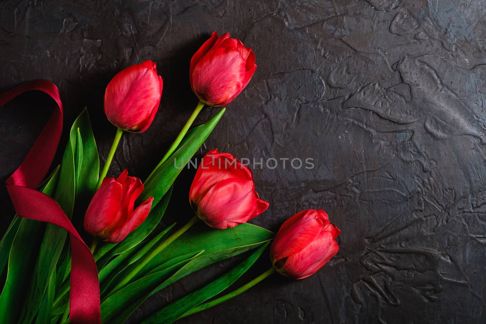 Red bunch of tulip flowers with red ribbon on textured black background, top view copy space