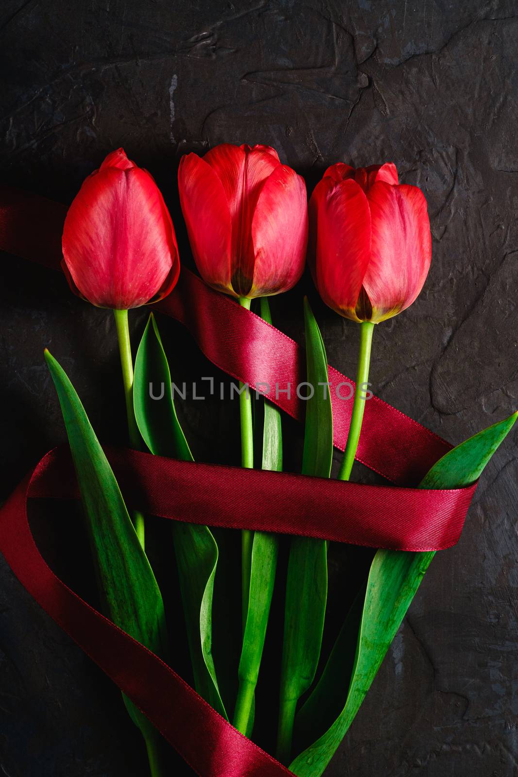 Red bunch of tulip flowers with red ribbon on textured black background, top view copy space