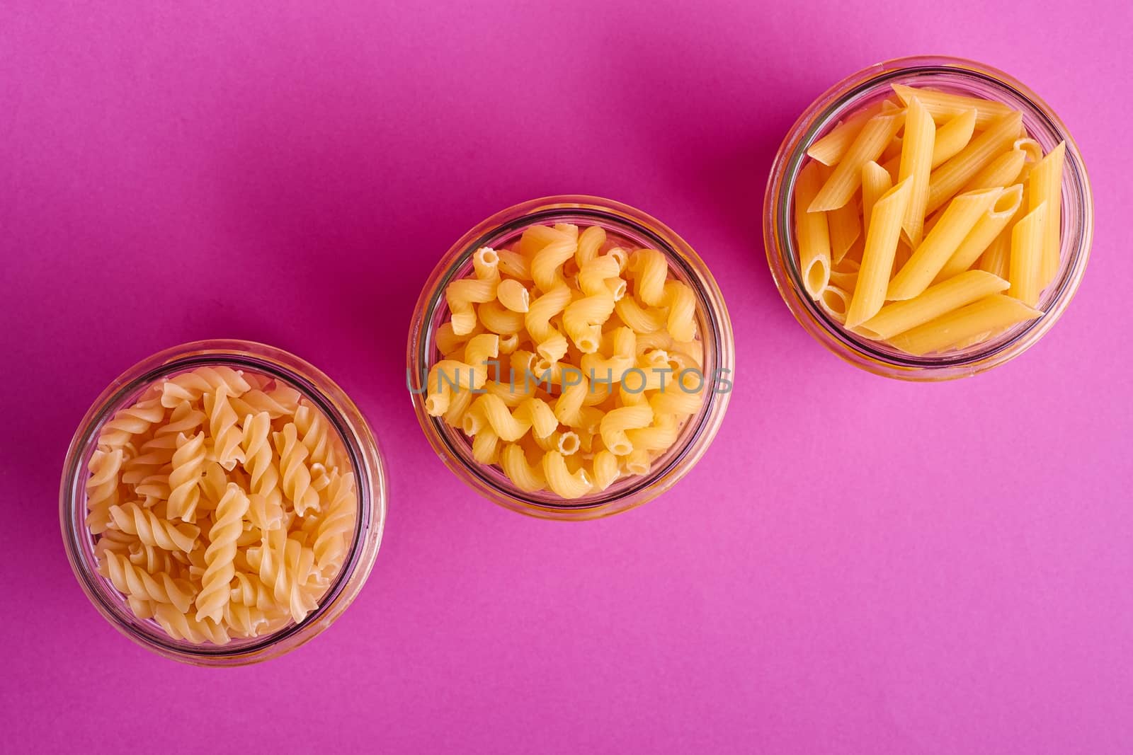 Three glass jars with variety of uncooked golden wheat pasta on minimal pink background, top view